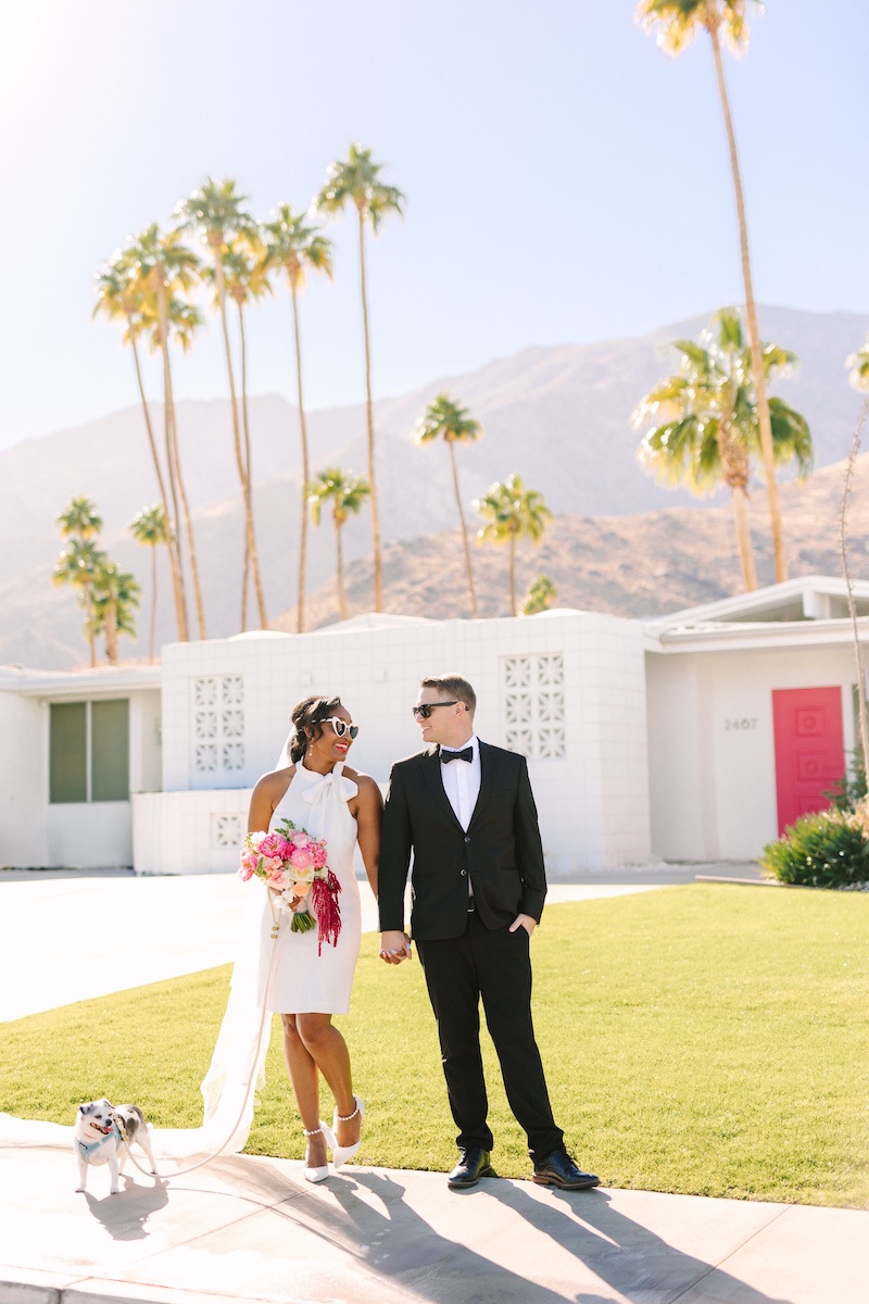 A couple in wedding attire holds hands and smiles outside a white mid-century modern house with palm trees and mountains in the background.