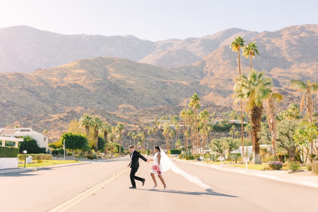 A couple in wedding attire holds hands and runs down a palm-lined street with mountains in the background under a clear sky.