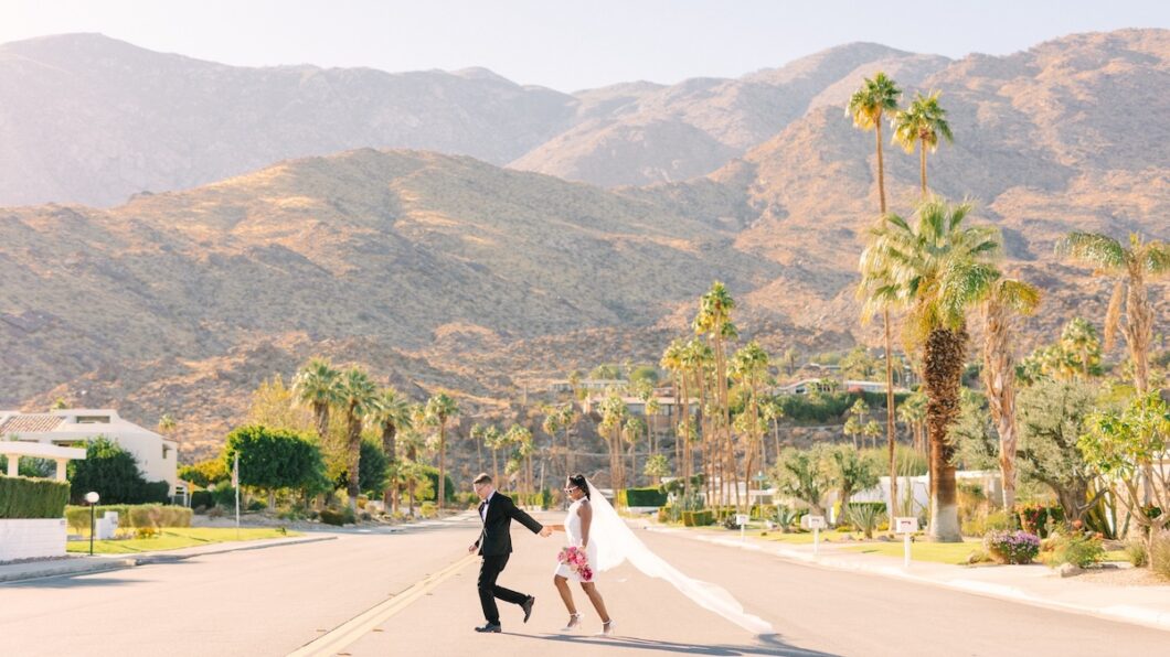 A couple in wedding attire holds hands and runs down a palm-lined street with mountains in the background under a clear sky.