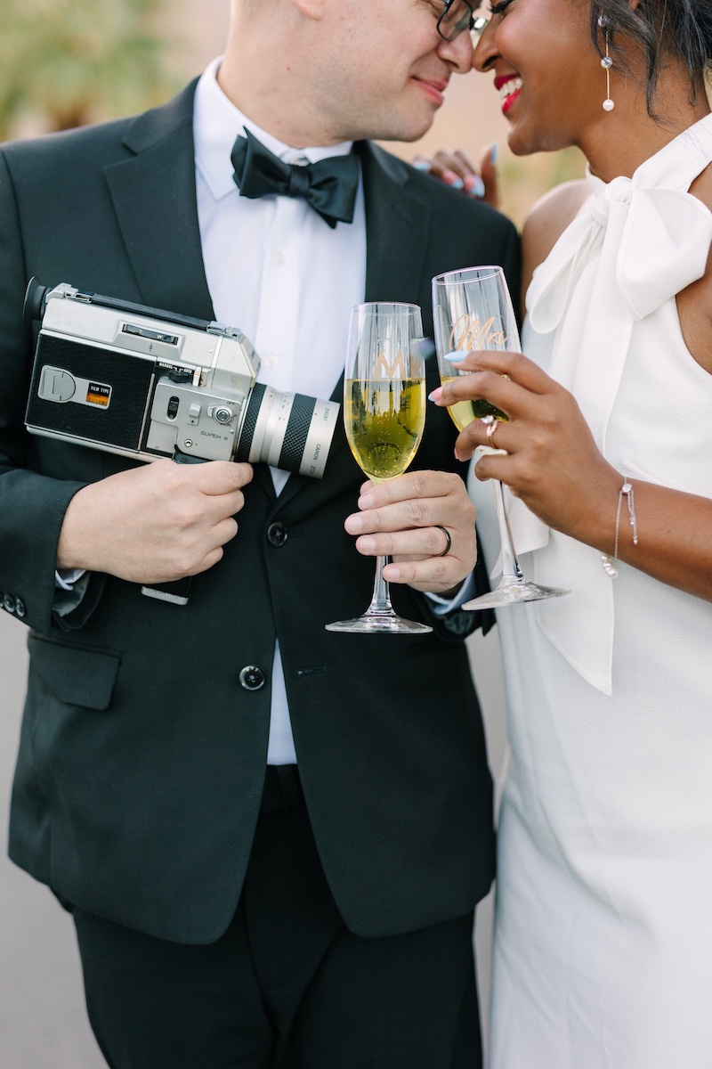 A couple dressed in formal attire holds champagne glasses and a vintage camera, standing close together and smiling.