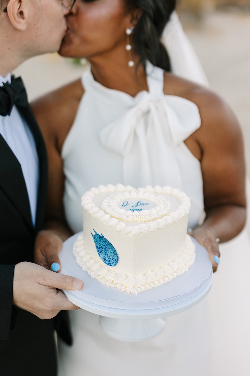 A bride and groom kiss while holding a small heart-shaped white cake with blue decorations on a white plate.