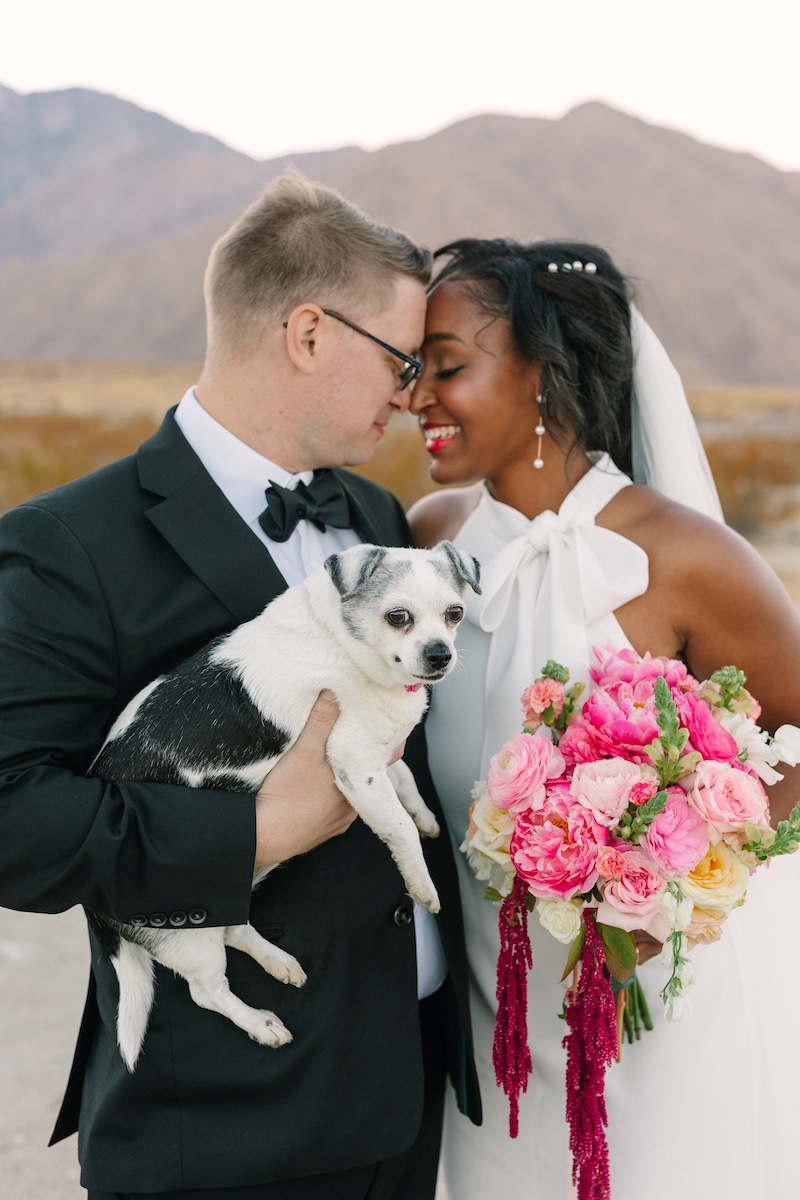 Bride and groom in formal attire stand close together outdoors, smiling with their foreheads touching. The groom holds a small dog, and the bride holds a bouquet of pink and white flowers.