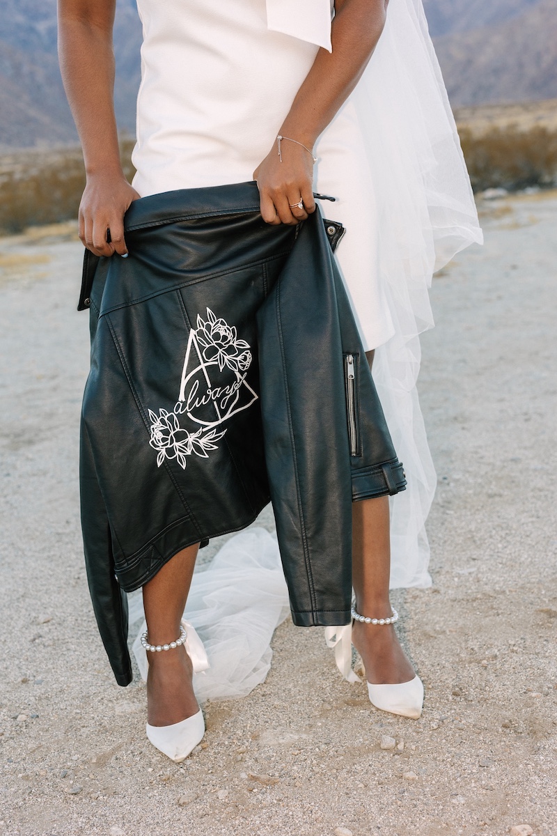 A person in a white dress and heels holds a black leather jacket with a geometric and floral design, standing outdoors on a sandy surface.