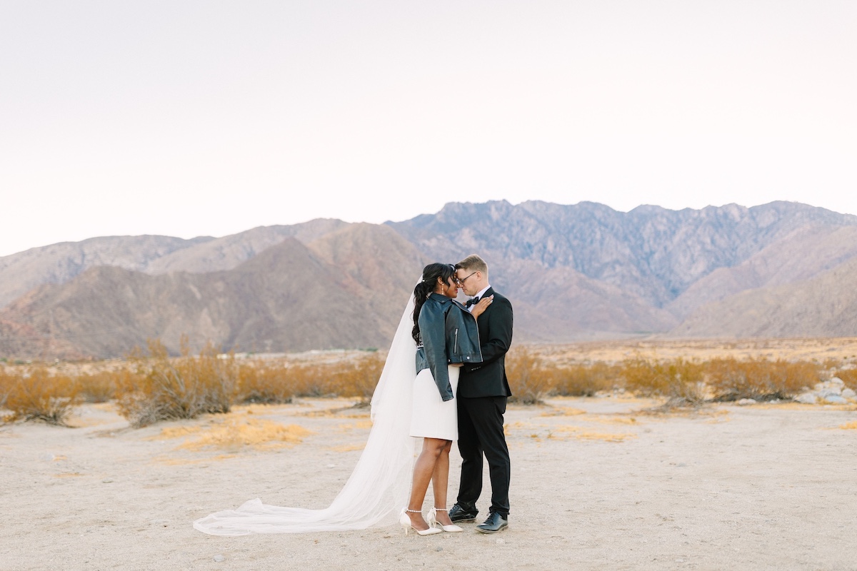 A bride and groom stand close together in a desert landscape, with mountains in the background. The bride wears a veil and a jacket, while the groom wears a suit.