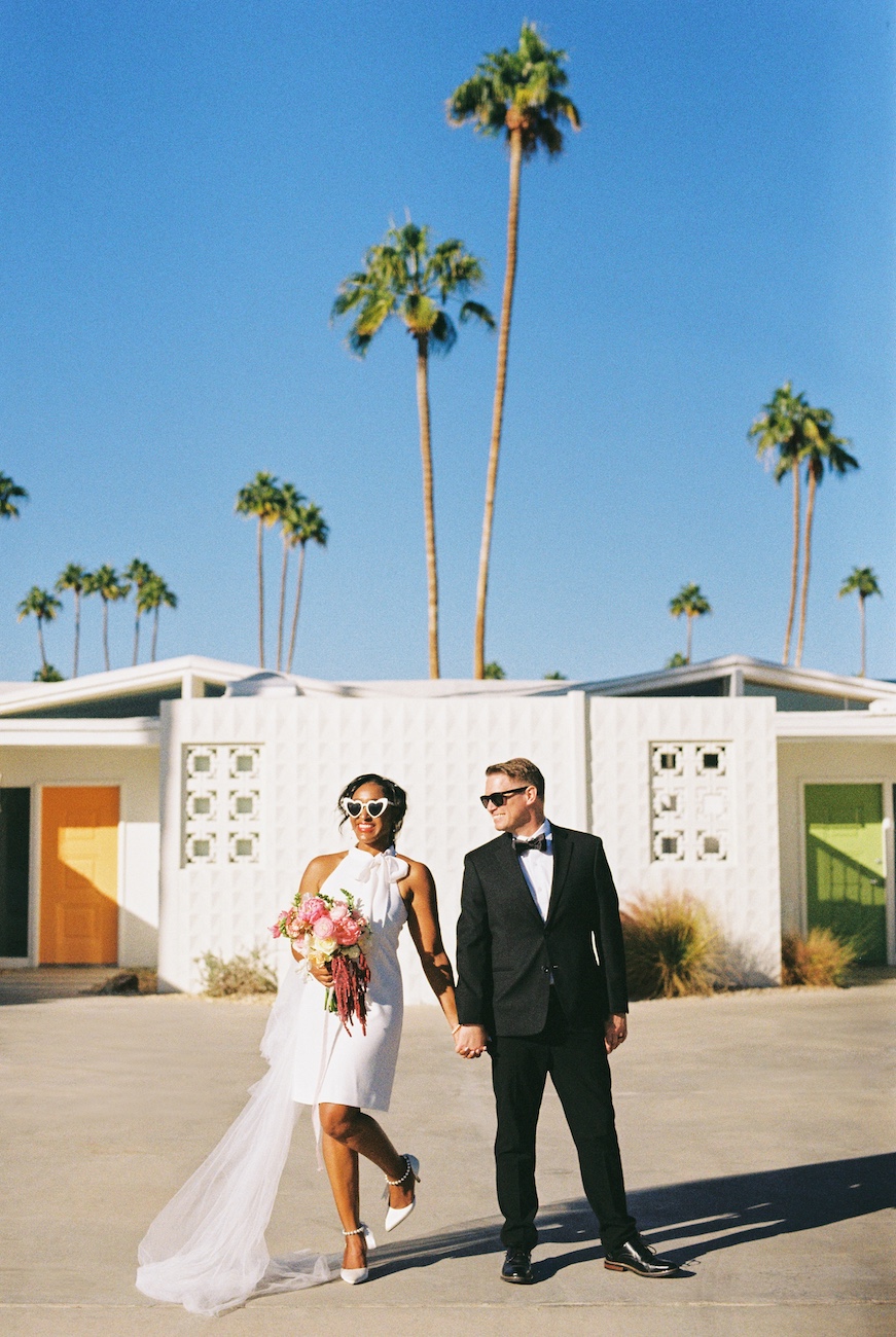 A bride and groom holding hands, walking outside on a sunny day, with palm trees and colorful doors in the background.