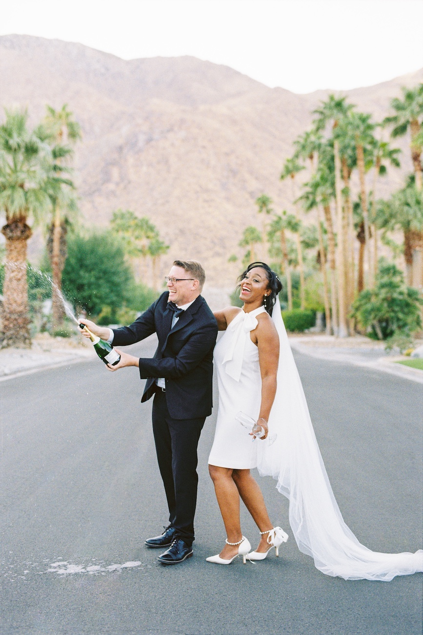 A couple in wedding attire stands on a palm-lined road; the groom pops a champagne bottle while the bride poses beside him, mountains visible in the background.