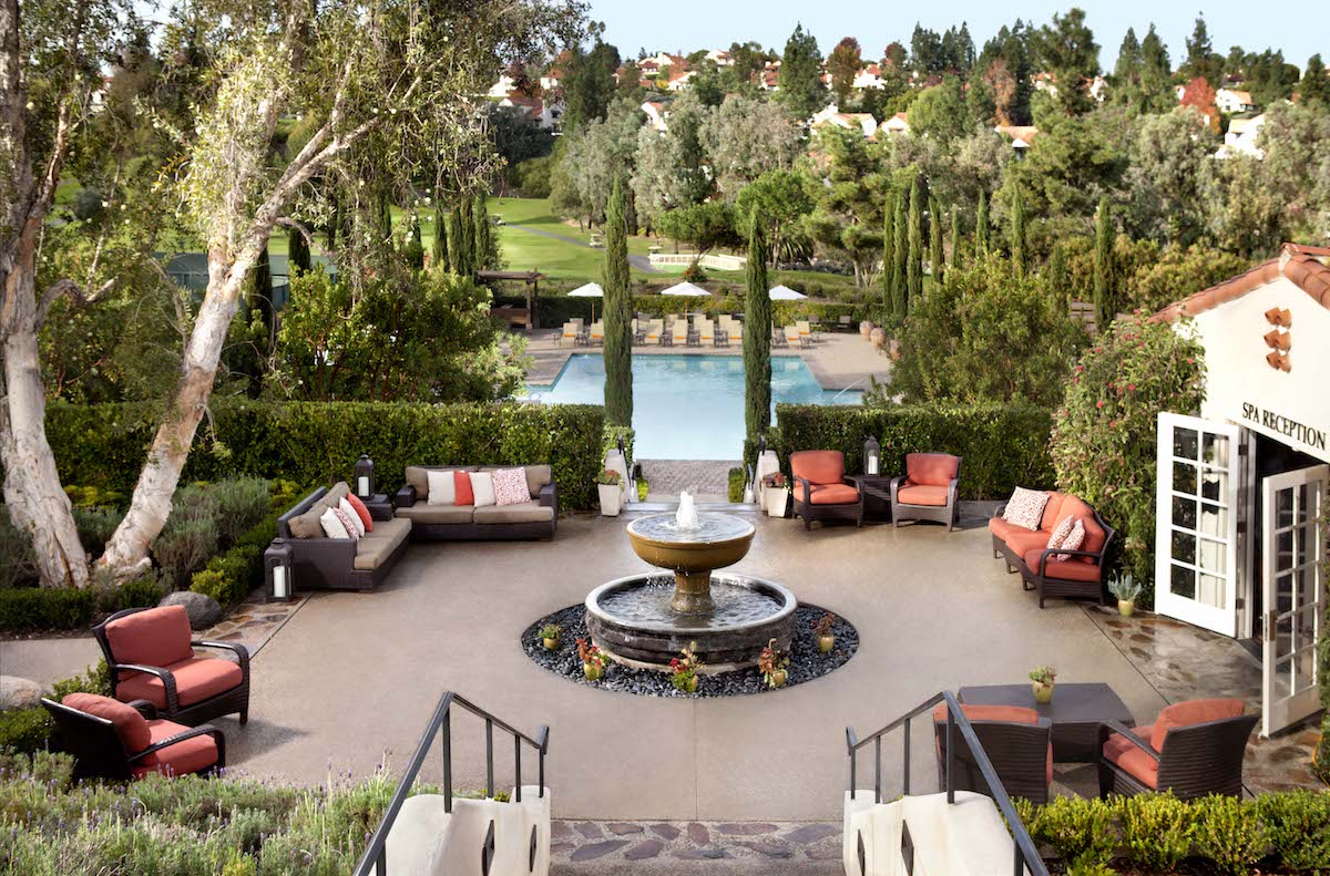 Outdoor patio with red cushioned seating around a central fountain, overlooking a pool and landscaped garden with tall trees and trimmed hedges.