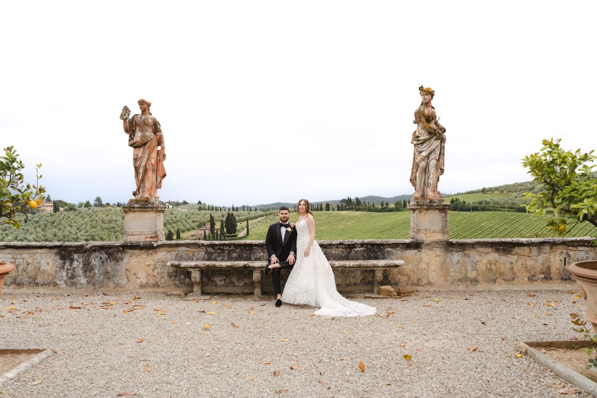 A bride and groom sit on a stone bench in an outdoor setting with statues and a scenic Tuscan landscape in the background.