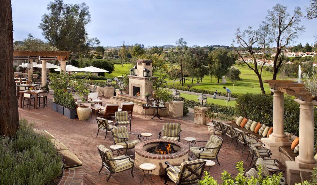 Outdoor patio with cushioned chairs around a fire pit, stone fireplace, and scenic view of a green lawn and trees in the background.