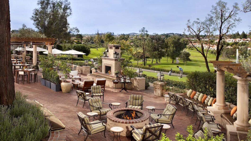 Outdoor patio with cushioned chairs around a fire pit, stone fireplace, and scenic view of a green lawn and trees in the background.