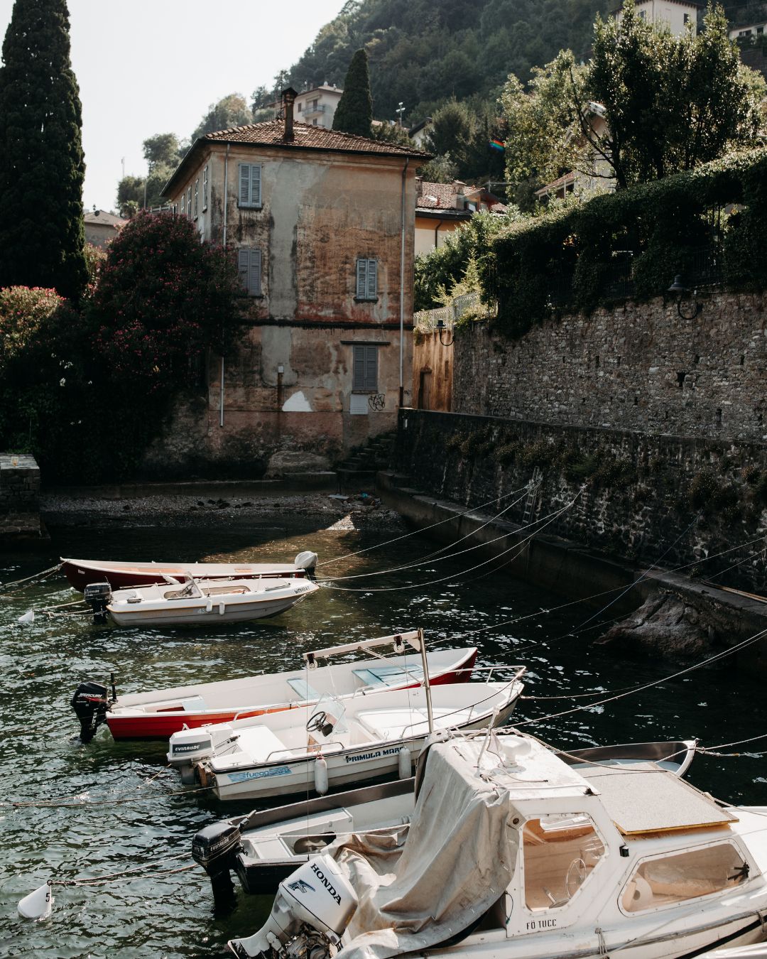 Four small boats are docked along a stone wall in a narrow canal beside an old, weathered building surrounded by greenery.
