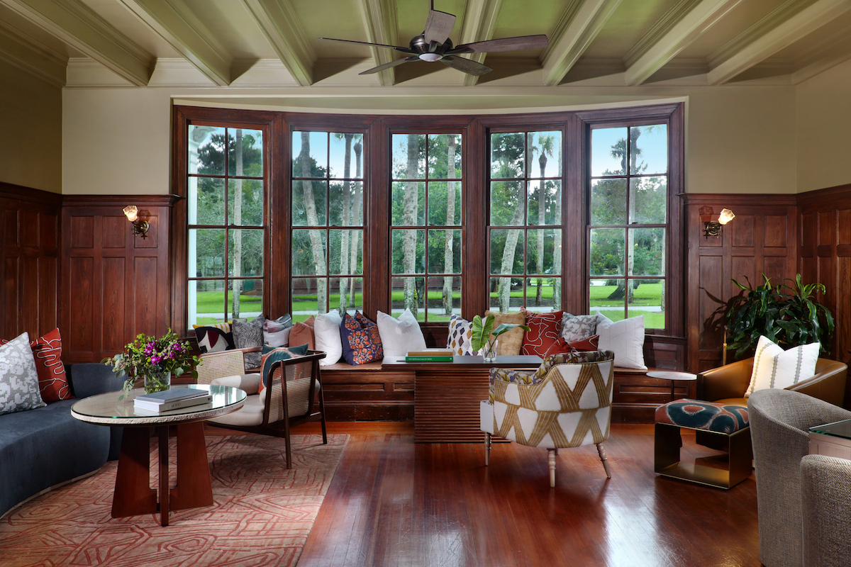 Spacious living room with wood paneling, large windows, built-in window seat with pillows, chairs, sofas, plants, and a ceiling fan.