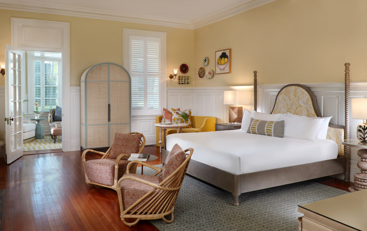 A spacious bedroom with a large bed, two wicker chairs, a small table, an armoire, hardwood floors, and a view into a sunroom.