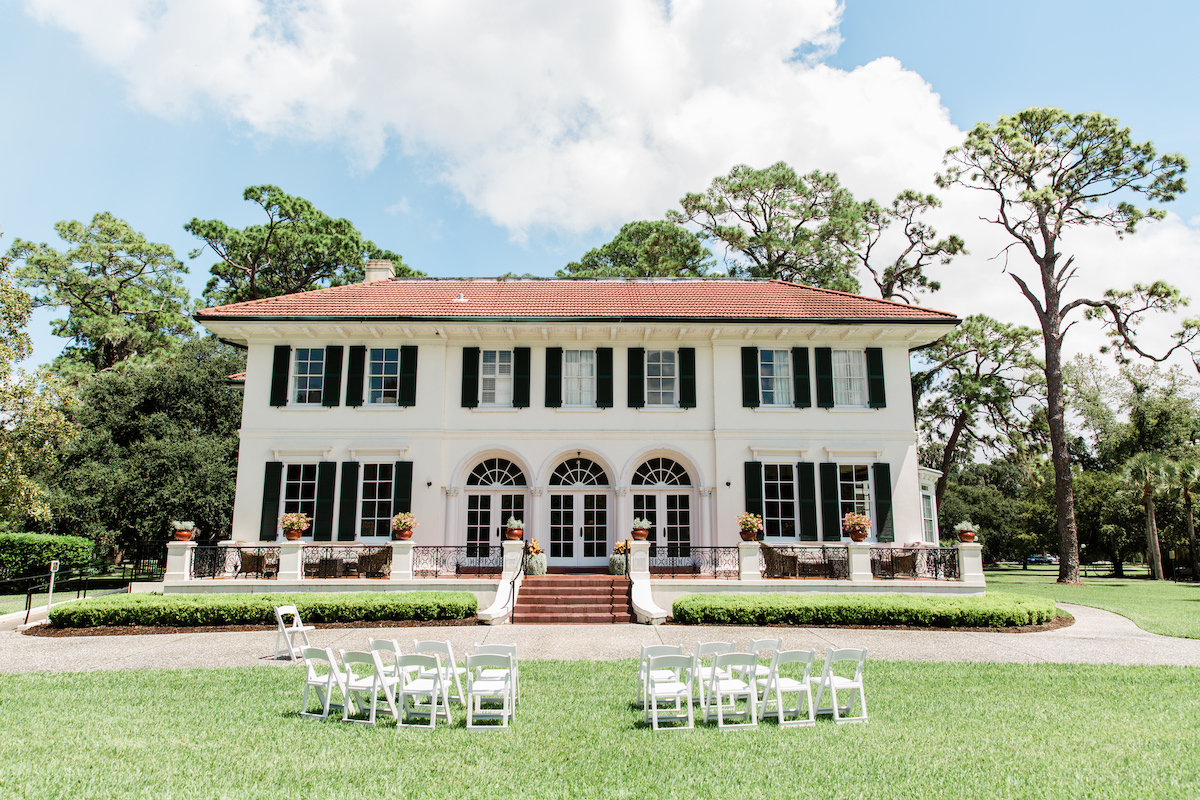 A large white two-story house with green shutters, a red roof, and arched windows, with white chairs arranged on the lawn in front.