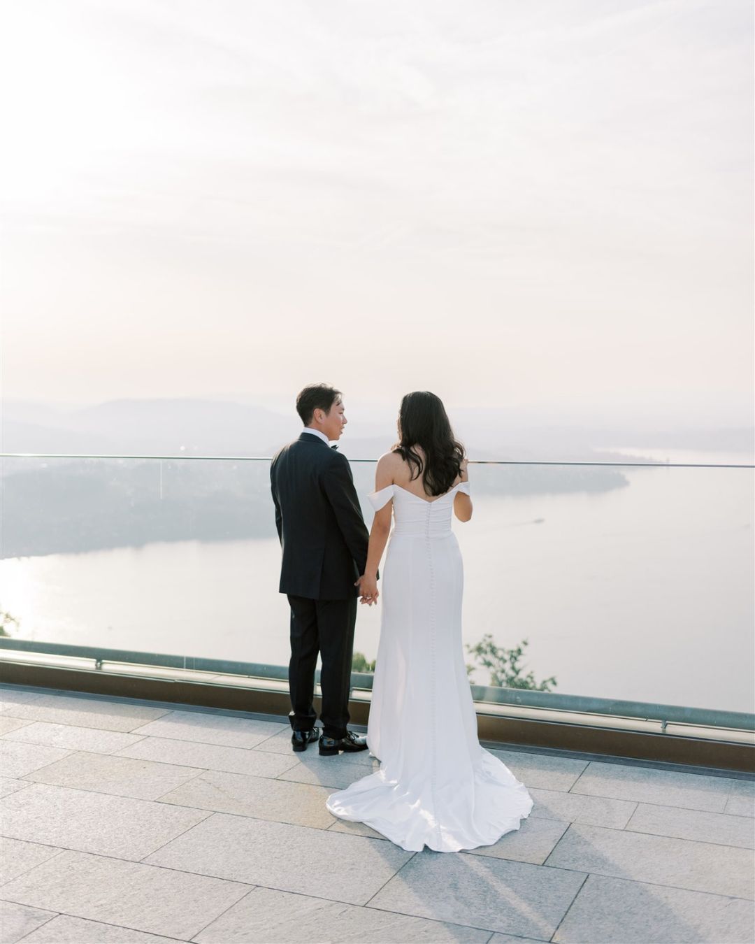 A couple dressed in formal attire stands side by side, holding hands and looking out over a calm body of water from a scenic overlook.