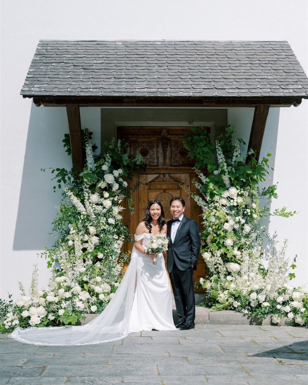 A bride and groom in formal attire stand together in front of a wooden door adorned with white and green floral arrangements.