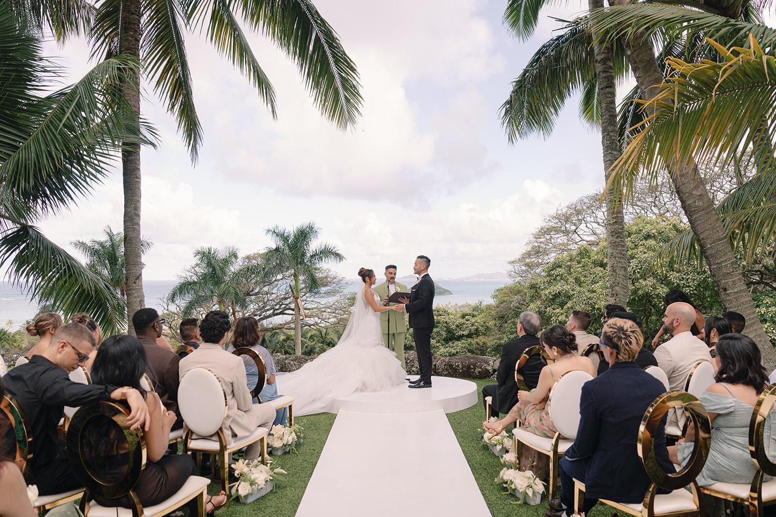 A bride and groom stand at an outdoor altar surrounded by guests, with palm trees and a view of the ocean in the background.