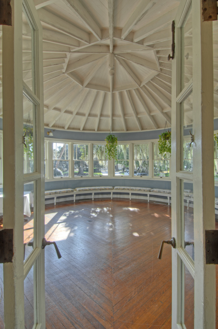 Double doors open to a circular room with a conical ceiling, wood floors, large windows, and hanging green plants.