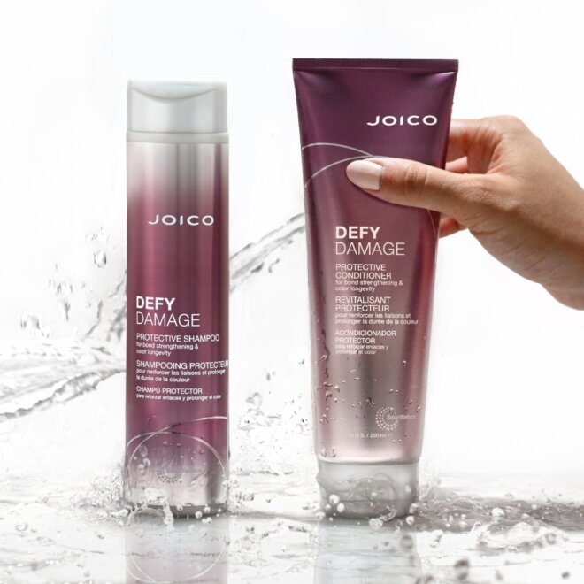 Two bottles of Joico Defy Damage hair products are shown; one bottle stands upright, while a hand holds the other against a white background with splashing water.
