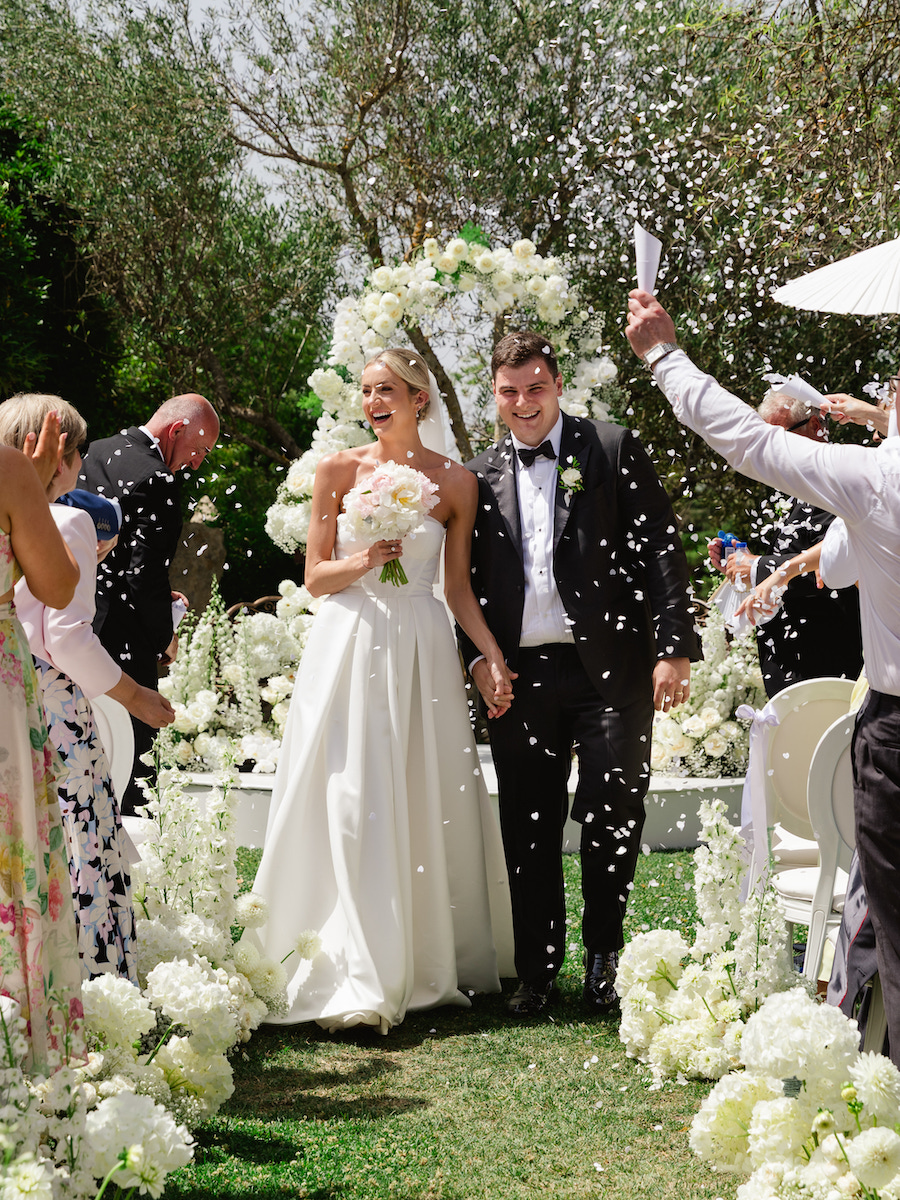 A bride and groom walk down an outdoor aisle, smiling as guests throw confetti. White flowers and greenery decorate the scene.