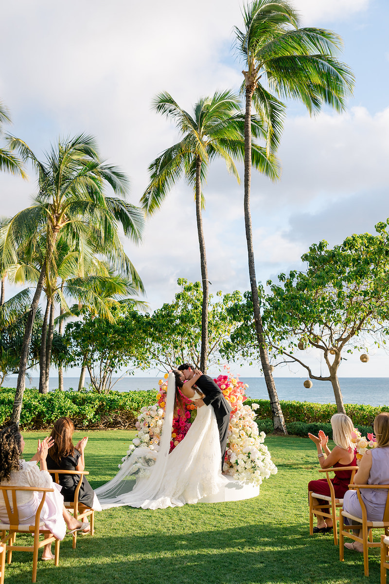 A bride and groom kiss at an outdoor wedding ceremony in front of palm trees, floral arrangements, and seated guests, with the ocean in the background.