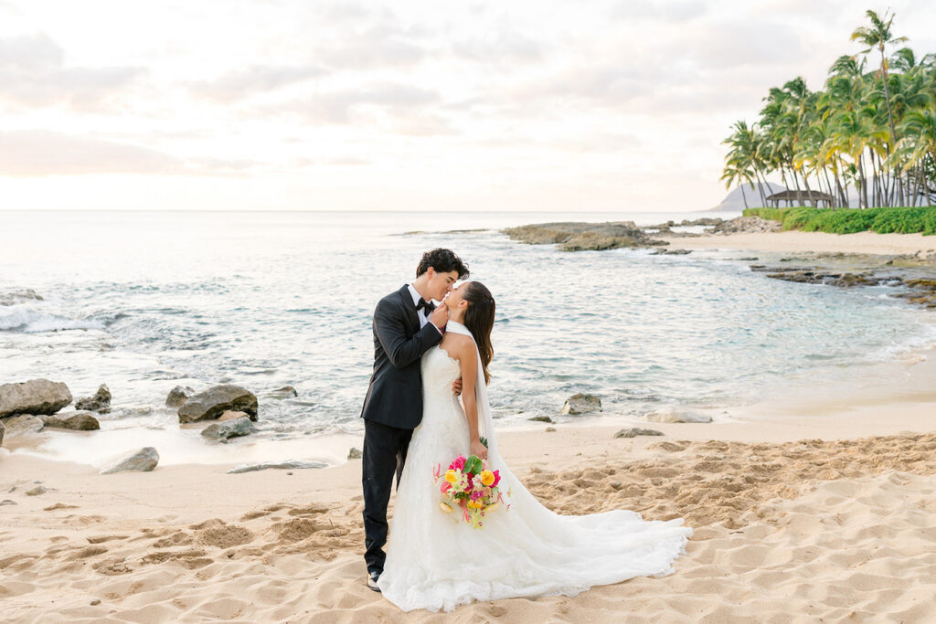 A bride and groom kiss on a sandy beach at sunset, with the ocean and palm trees in the background. The bride holds a colorful bouquet.