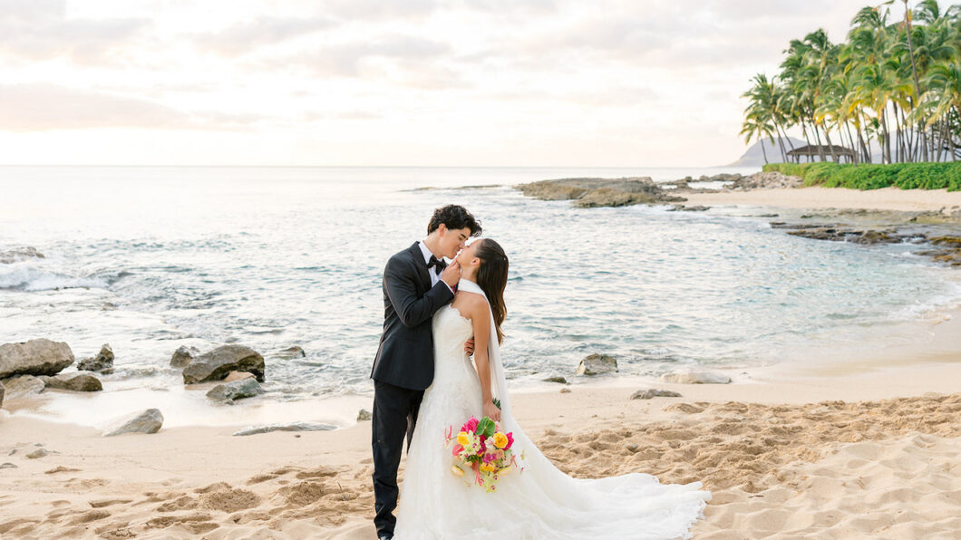 A bride and groom kiss on a sandy beach at sunset, with the ocean and palm trees in the background. The bride holds a colorful bouquet.