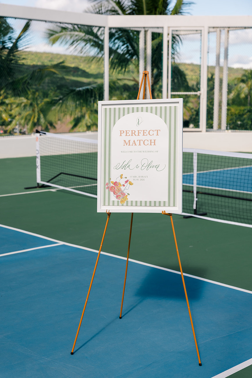A sign on an easel reading “Perfect Match” stands on a tennis court, with palm trees and a net visible in the background.