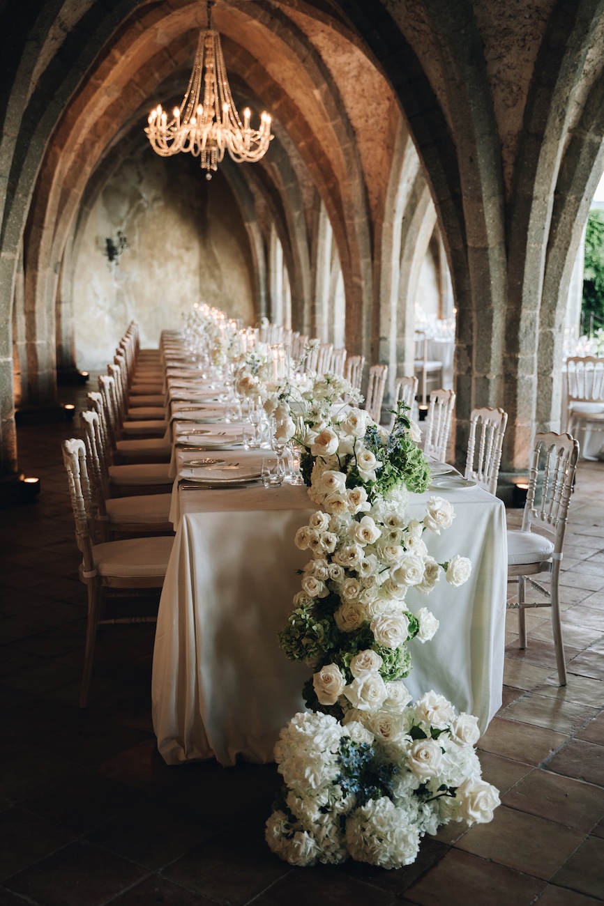 Long banquet table with white tablecloth set for a formal event, decorated with cascading white flowers in an arched stone hall with a chandelier.