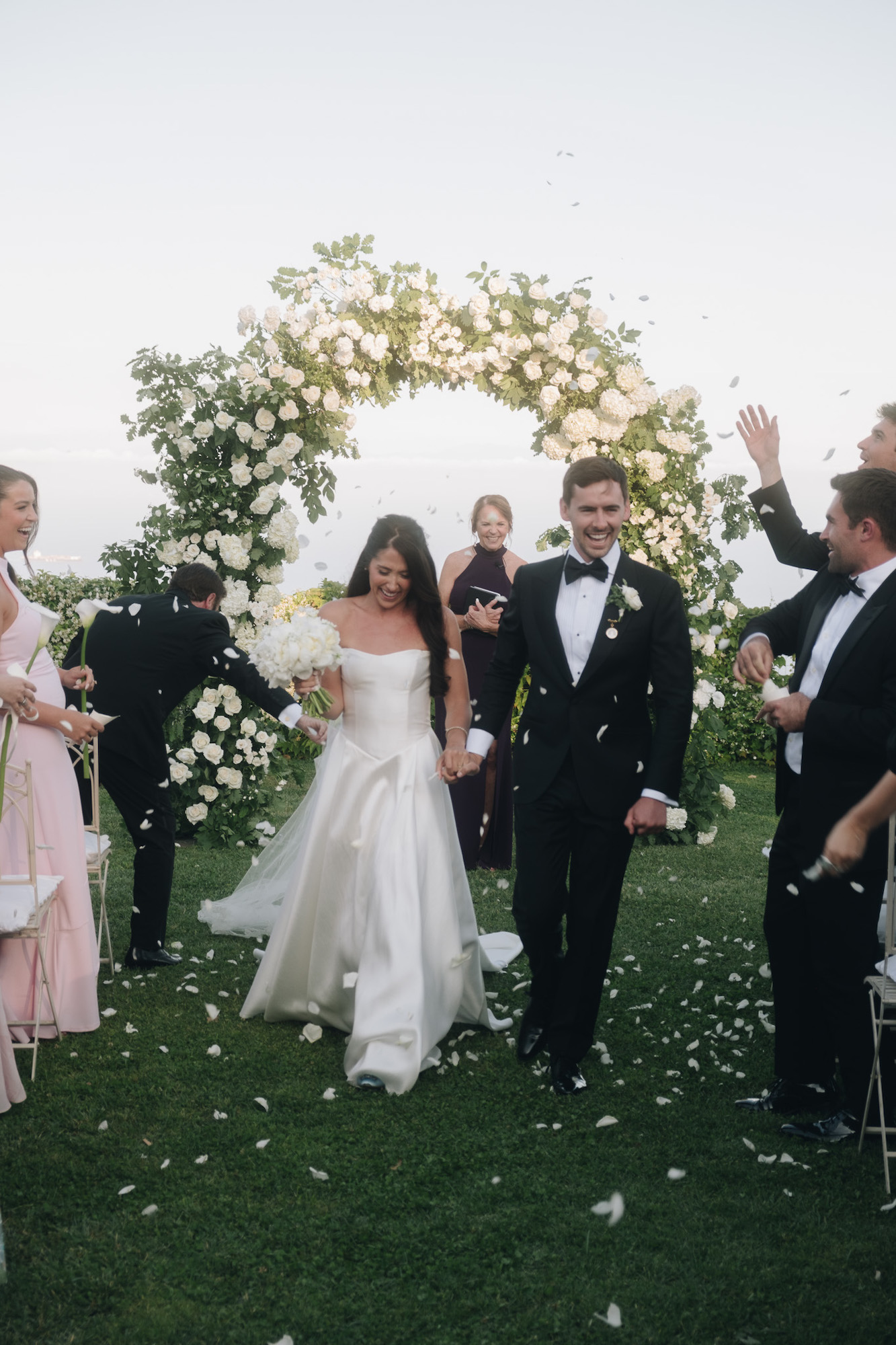 A bride and groom walk down the aisle outdoors, surrounded by guests and a circular floral arch. Guests toss flower petals as the couple smiles.