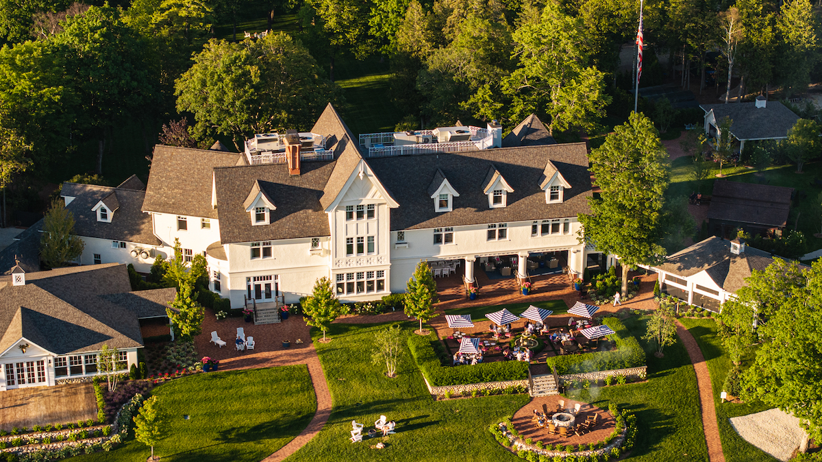 A large, light-colored manor-style building with multiple gables surrounded by green lawns, gardens, patios, and outdoor dining areas, viewed from above.