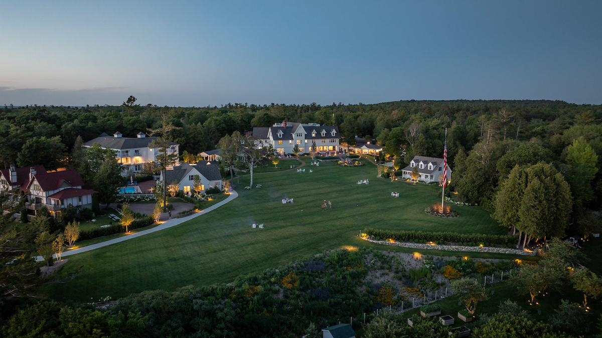 A large grassy lawn is surrounded by several buildings and trees at dusk, with outdoor lights illuminating pathways and scattered groups of people.