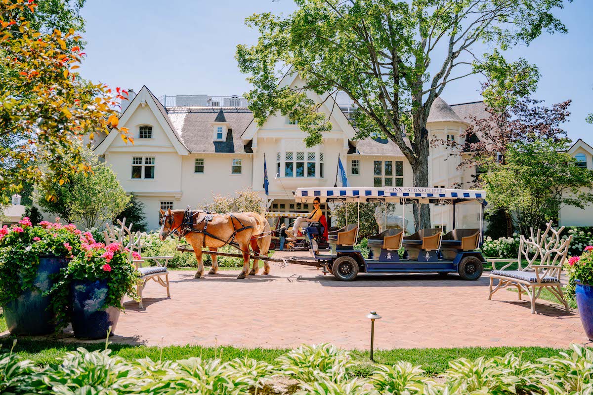 A horse-drawn carriage with a striped canopy is parked on a brick path in front of a large white house with lush landscaping.