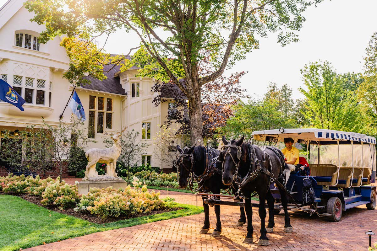 A horse-drawn carriage is parked on a brick path in front of a large house with flags and a lion statue, surrounded by greenery.