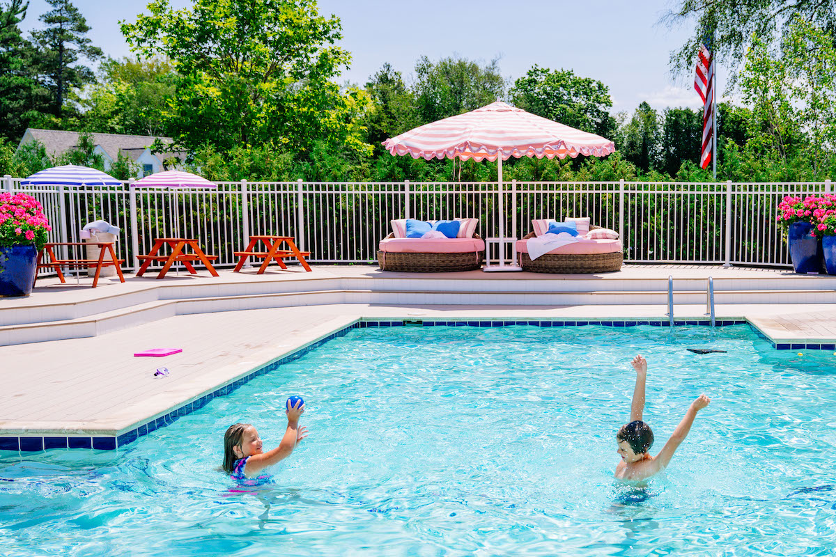 Two children play in a backyard swimming pool with lounge chairs, pink umbrellas, and flowers arranged on the pool deck in the background.