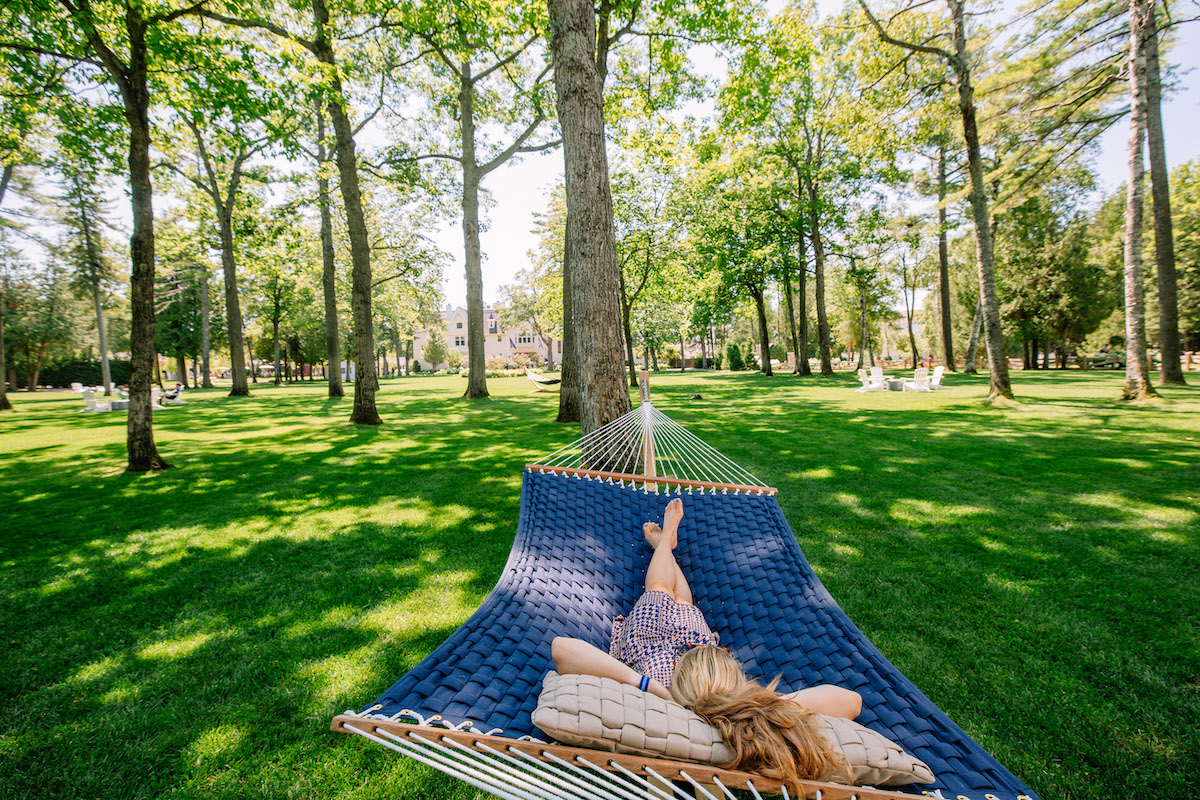 A person lies on a blue hammock in a grassy park surrounded by tall trees on a sunny day.