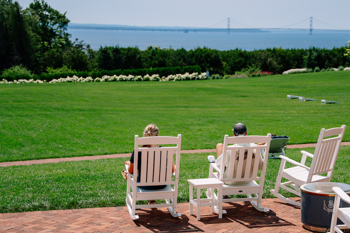 Two people sit on white rocking chairs facing a large grassy lawn with water and a bridge visible in the distance under a clear sky.