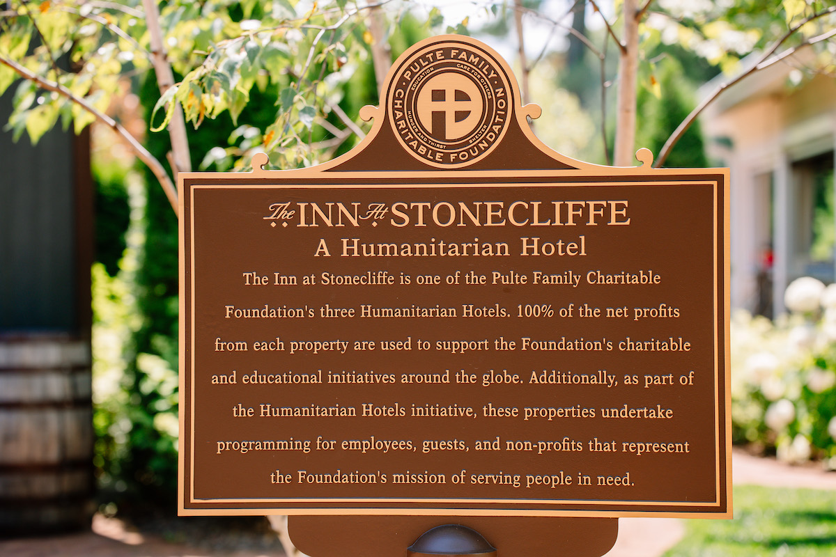 Bronze plaque reads: “The Inn at Stonecliffe, A Humanitarian Hotel,” describing charitable support from hotel profits for global humanitarian and educational initiatives. Trees and greenery are visible behind.