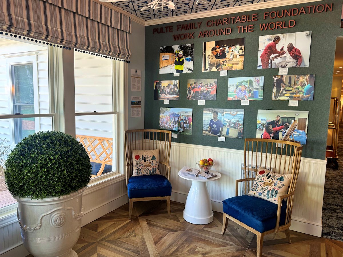 Two wooden chairs with blue cushions sit beside a small white table and a potted plant, near a wall displaying photos and text about a charitable foundation.