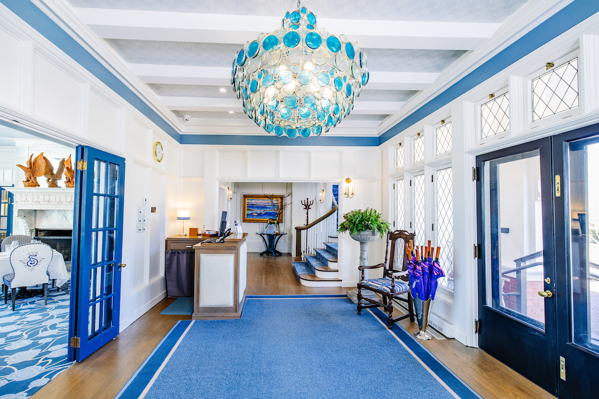 Bright hotel lobby with blue and white decor, large blue glass chandelier, blue doors, reception desk, and a staircase in the background.