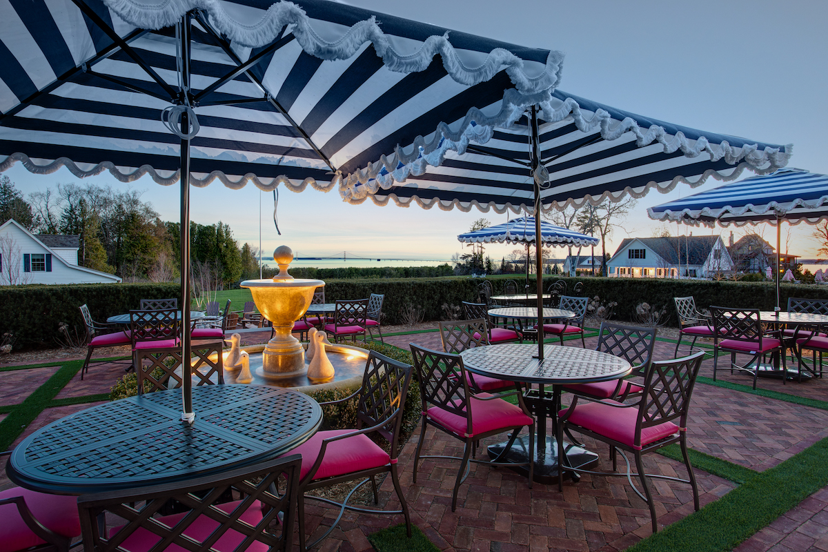 Outdoor patio with metal tables and pink cushioned chairs under striped umbrellas, featuring a central water fountain and a scenic landscape in the background at sunset.
