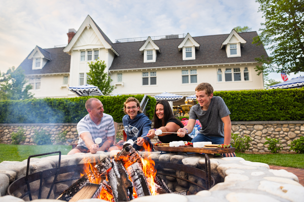 Four people sit around a stone fire pit roasting marshmallows in front of a large white house with a dark roof and hedges.
