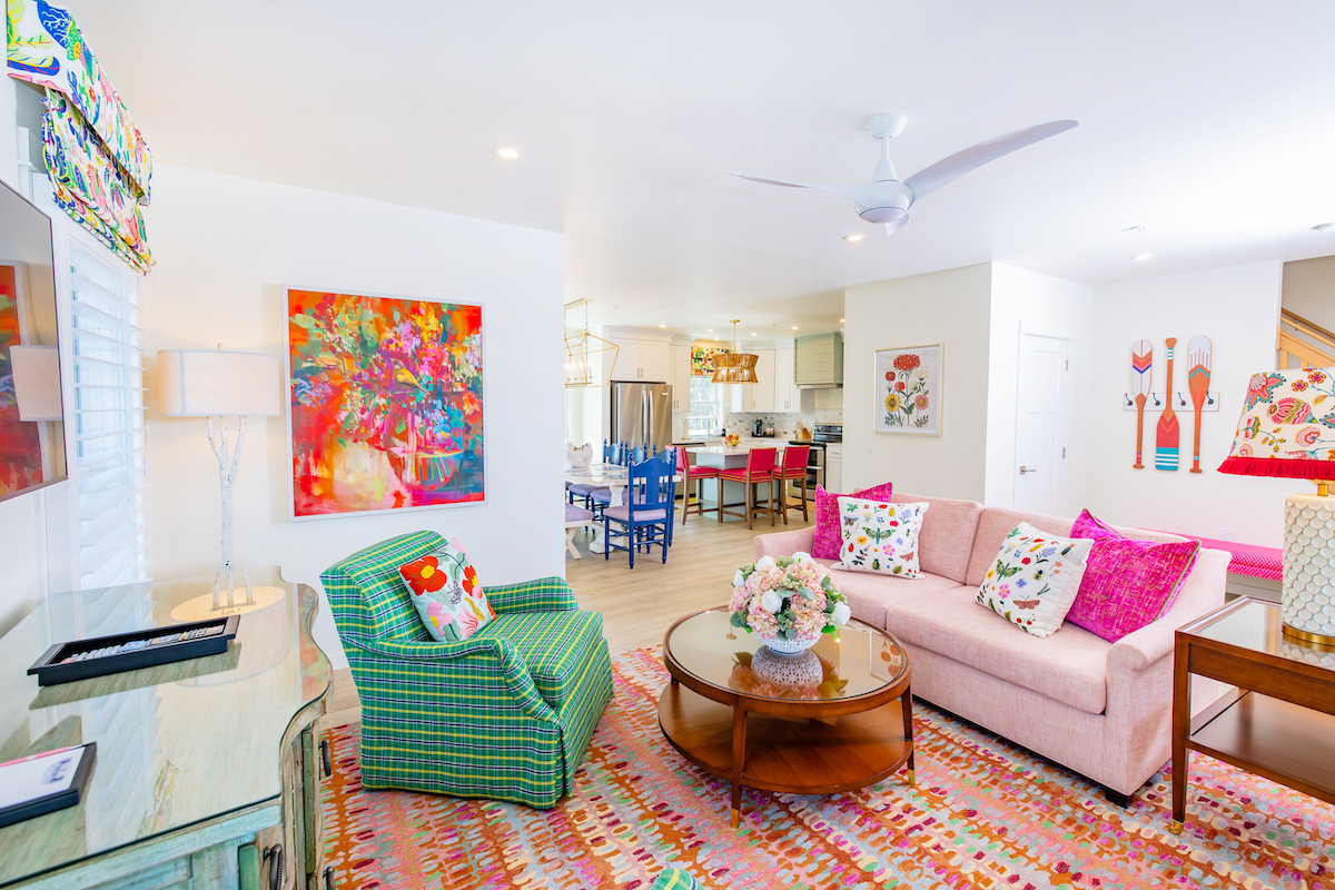 Bright, colorful living room with patterned furniture, a pink sofa, a green chair, vibrant artwork, and an open layout leading to a modern kitchen and dining area.
