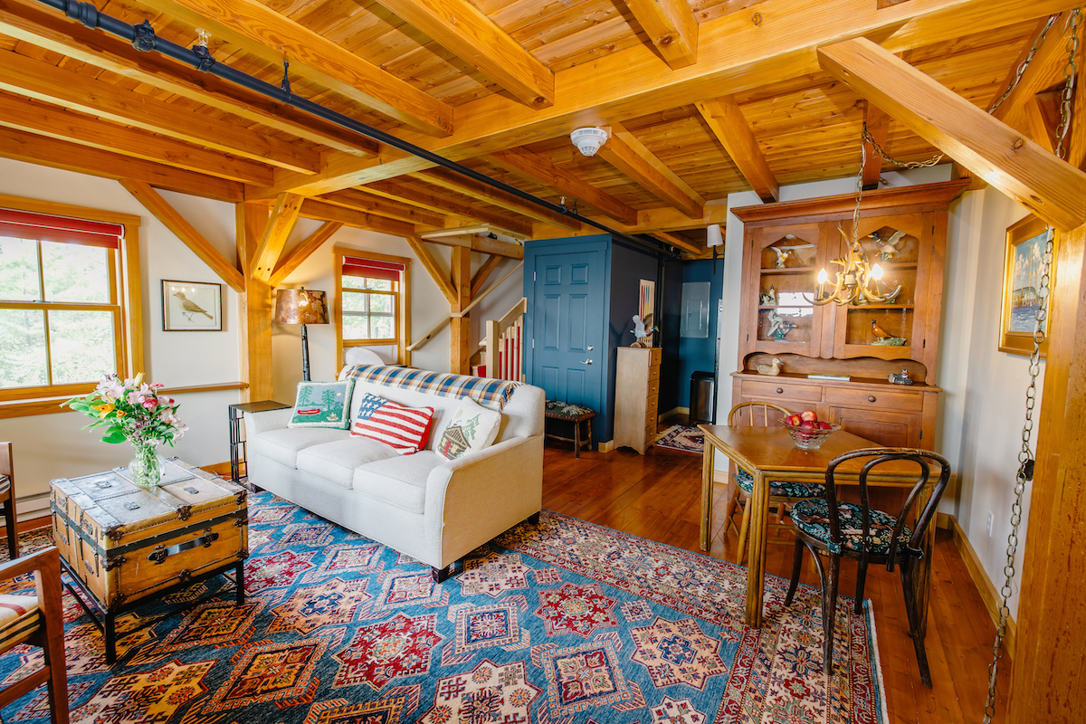 A cozy living room with exposed wooden beams, a white sofa, patterned rug, wooden dining table, blue accent wall, and bright natural light from multiple windows.