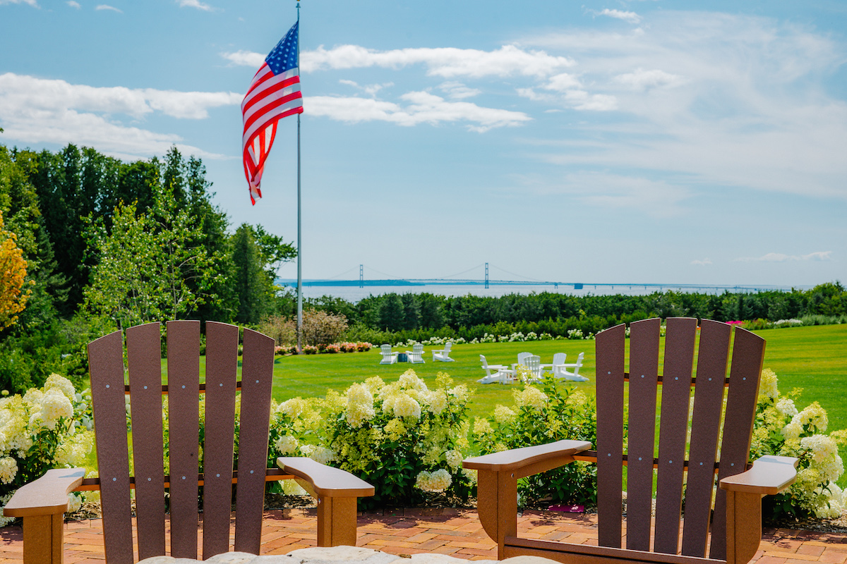 Two wooden Adirondack chairs face a grassy lawn with white flowers, a large American flag, and a distant view of water and a bridge under a blue sky with clouds.