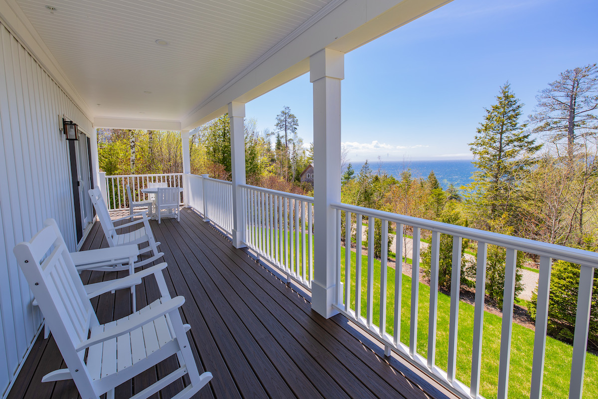 White rocking chairs on a covered porch with dark wood flooring, white railing, and a scenic view of trees, lawn, and water under a clear sky.
