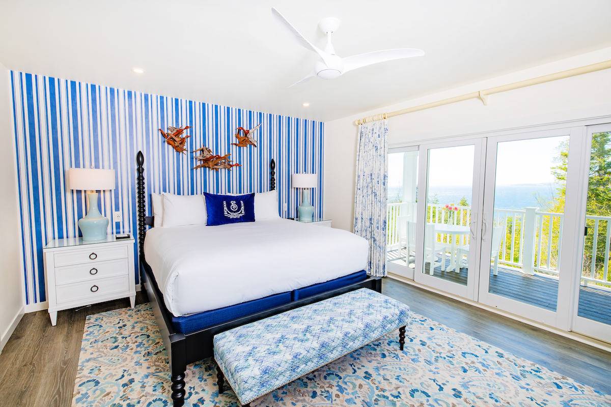 A bright bedroom with a large bed, striped blue accent wall, two nightstands, bench at the foot of the bed, and glass doors leading to a balcony with a scenic view.