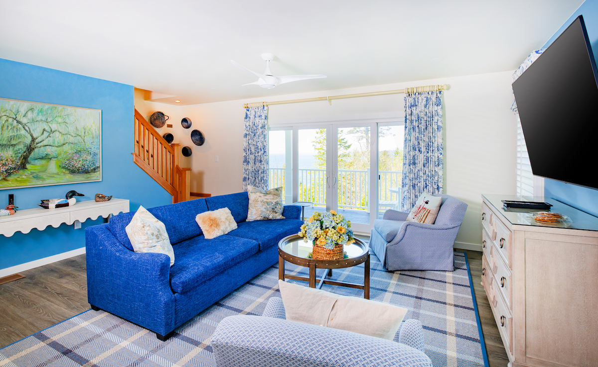Bright living room with blue sofa, two armchairs, plaid rug, wall-mounted TV, large windows with curtains, a staircase, and decorative plates on a blue accent wall.