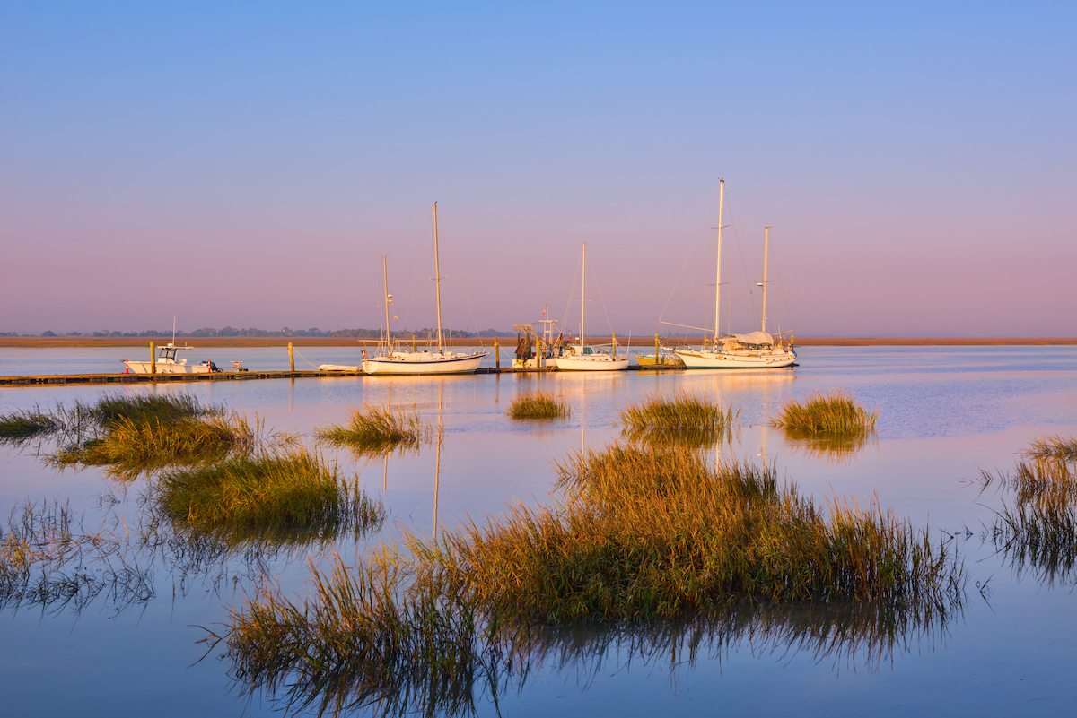 Several sailboats and yachts are docked at a pier on calm water, surrounded by patches of marsh grass, under a clear sky with soft pink and blue hues.