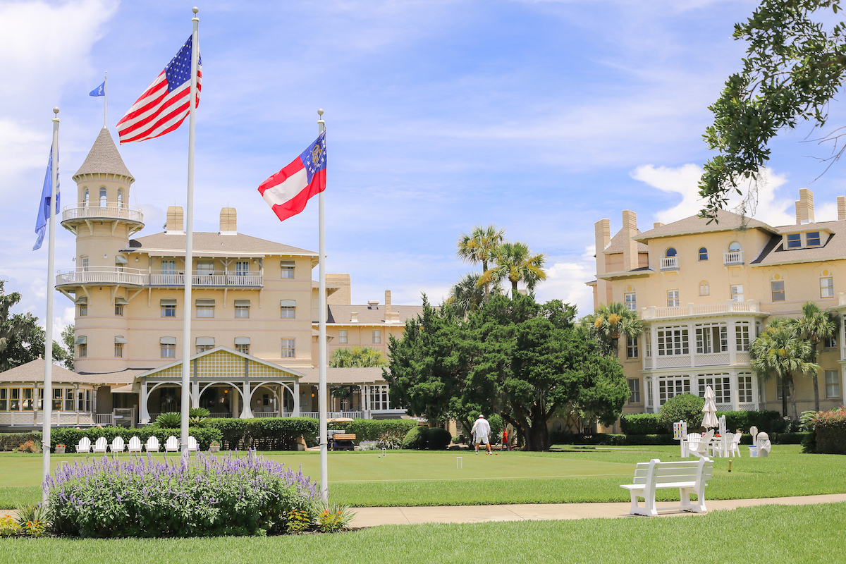 Historic beige hotel buildings with three flags in front, a green lawn, white benches, and a few people in the background under a blue sky with clouds.